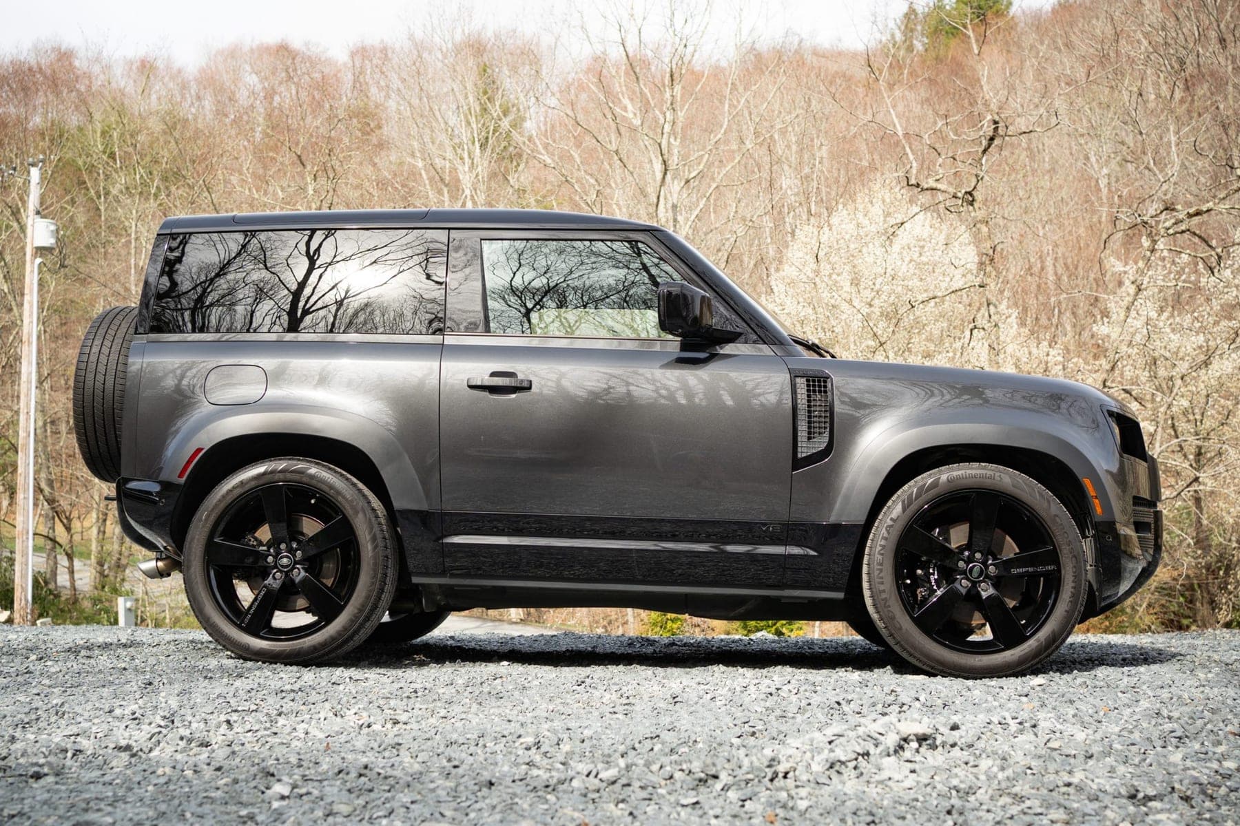 Grey Land Rover Defender 90 in side profile on gravel, with the spare wheel mounted on the tailgate