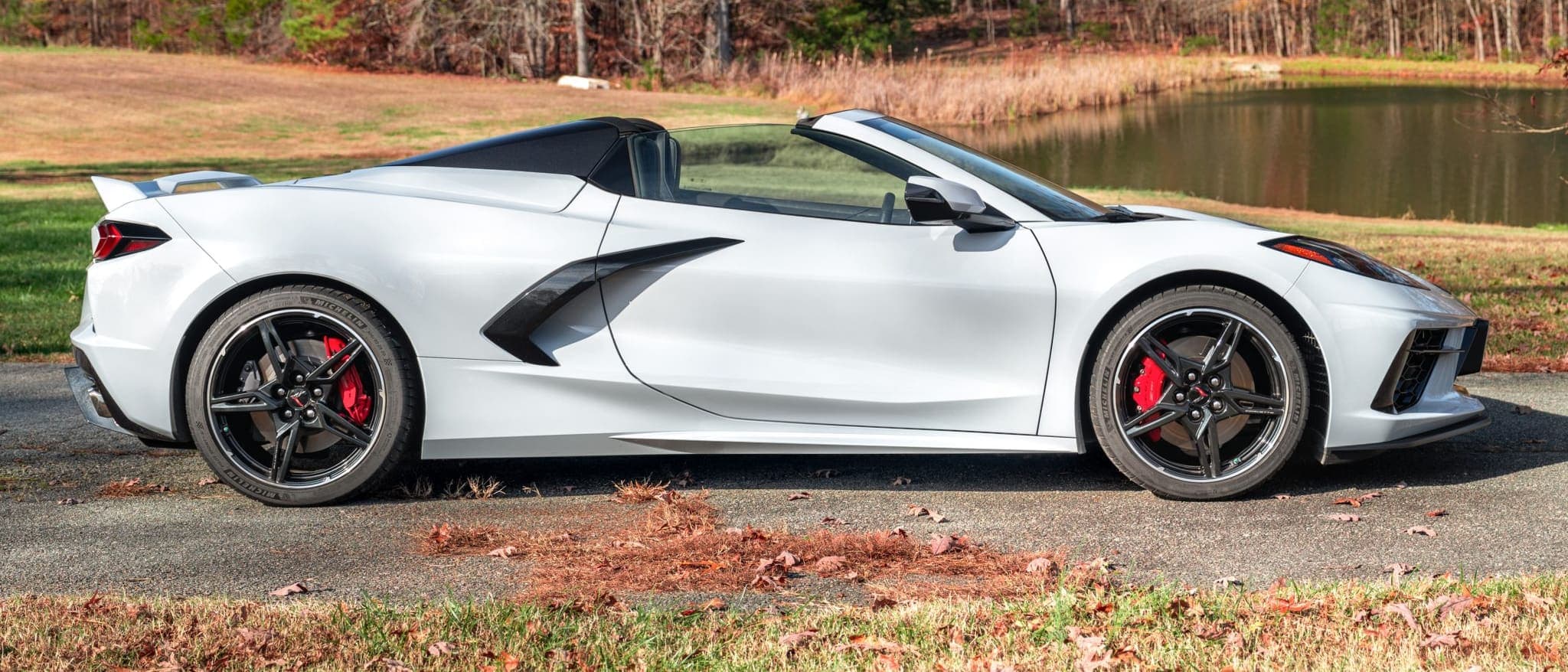 Side profile of a white Corvette Stingray convertible parked beside a pond in late autumn light