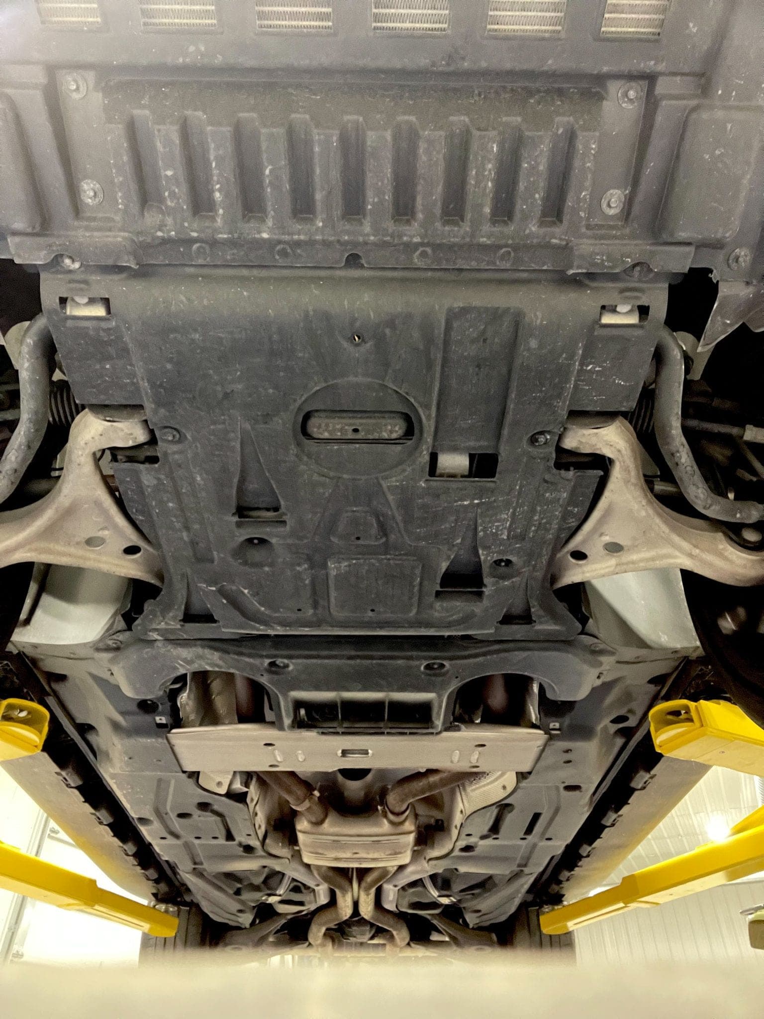 Underbody of a Mercedes-AMG GLS 63 raised on a two-post lift, the yellow lift arms in frame, showing the transmission tunnel, exhaust and front subframe