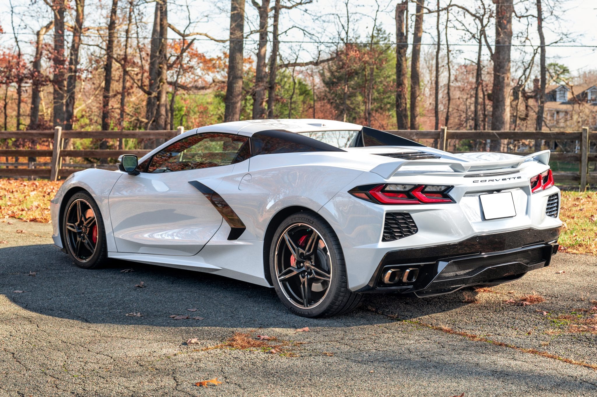 White Chevrolet Corvette Stingray convertible photographed from the rear three-quarter with the roof stowed, against bare autumn oaks — from a Carviondealer listing